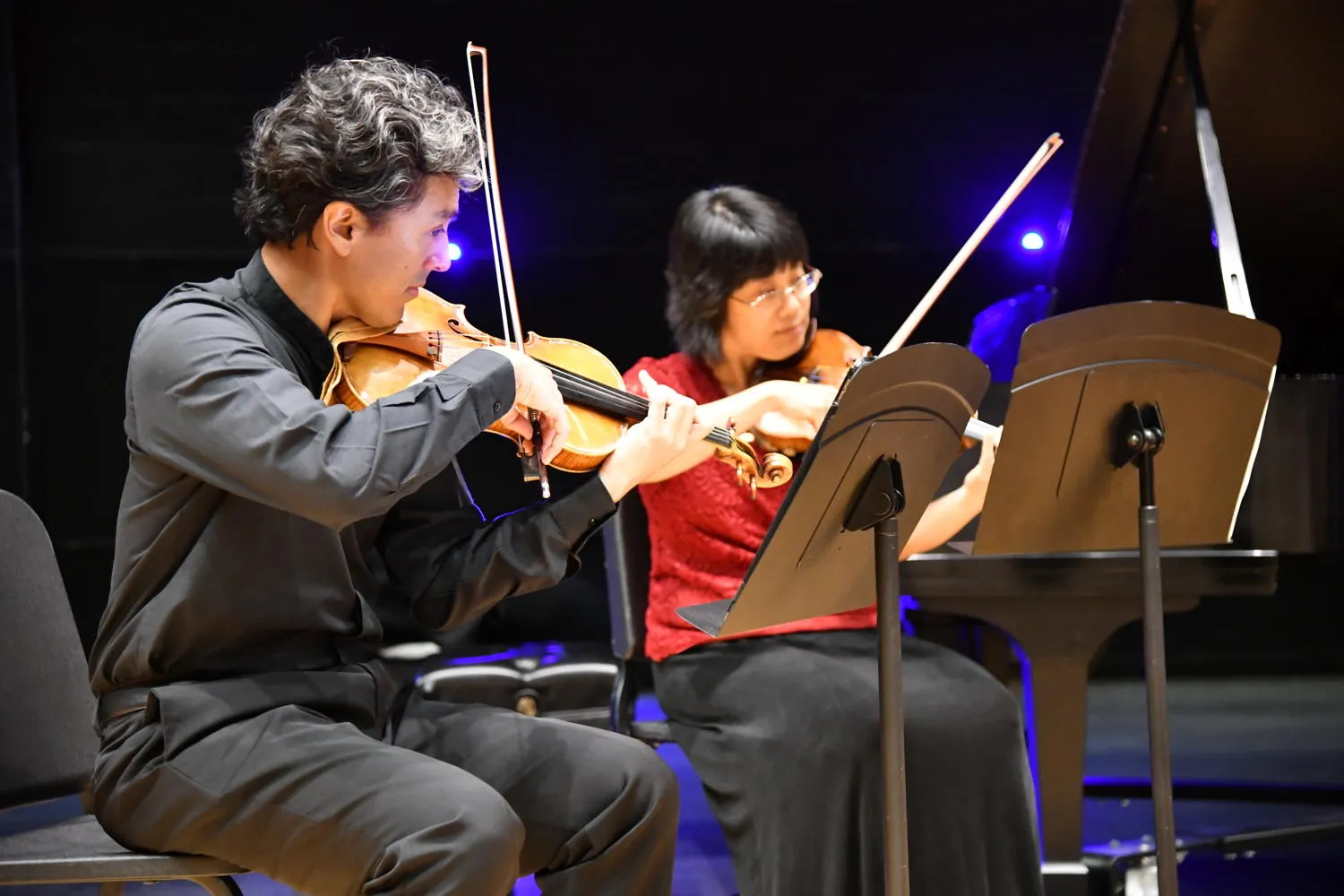 Two string players, one in black and one in red, playing side by side beside a grand piano