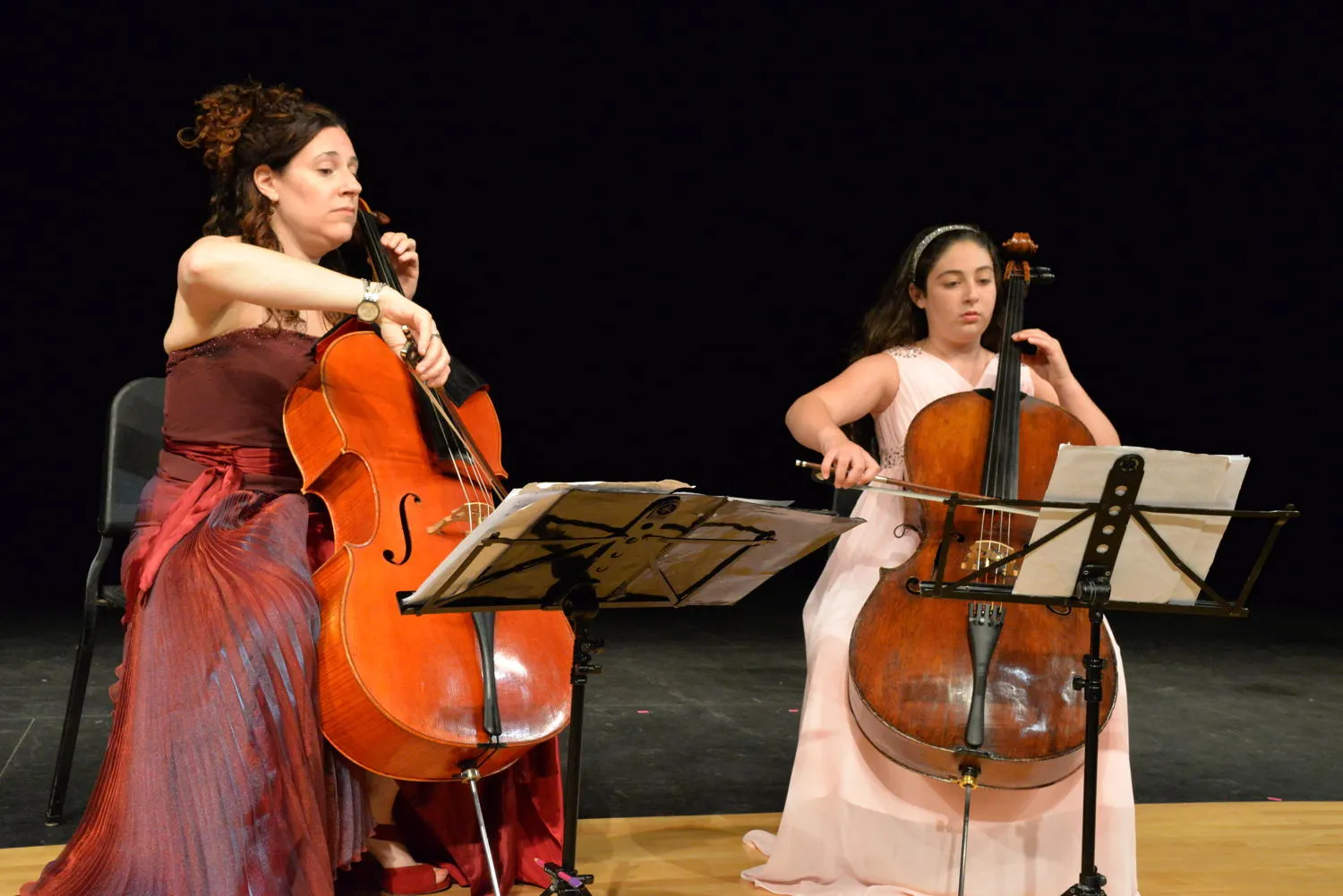 Two cellists, one in a red gown and a young player in a pink gown, playing side by side