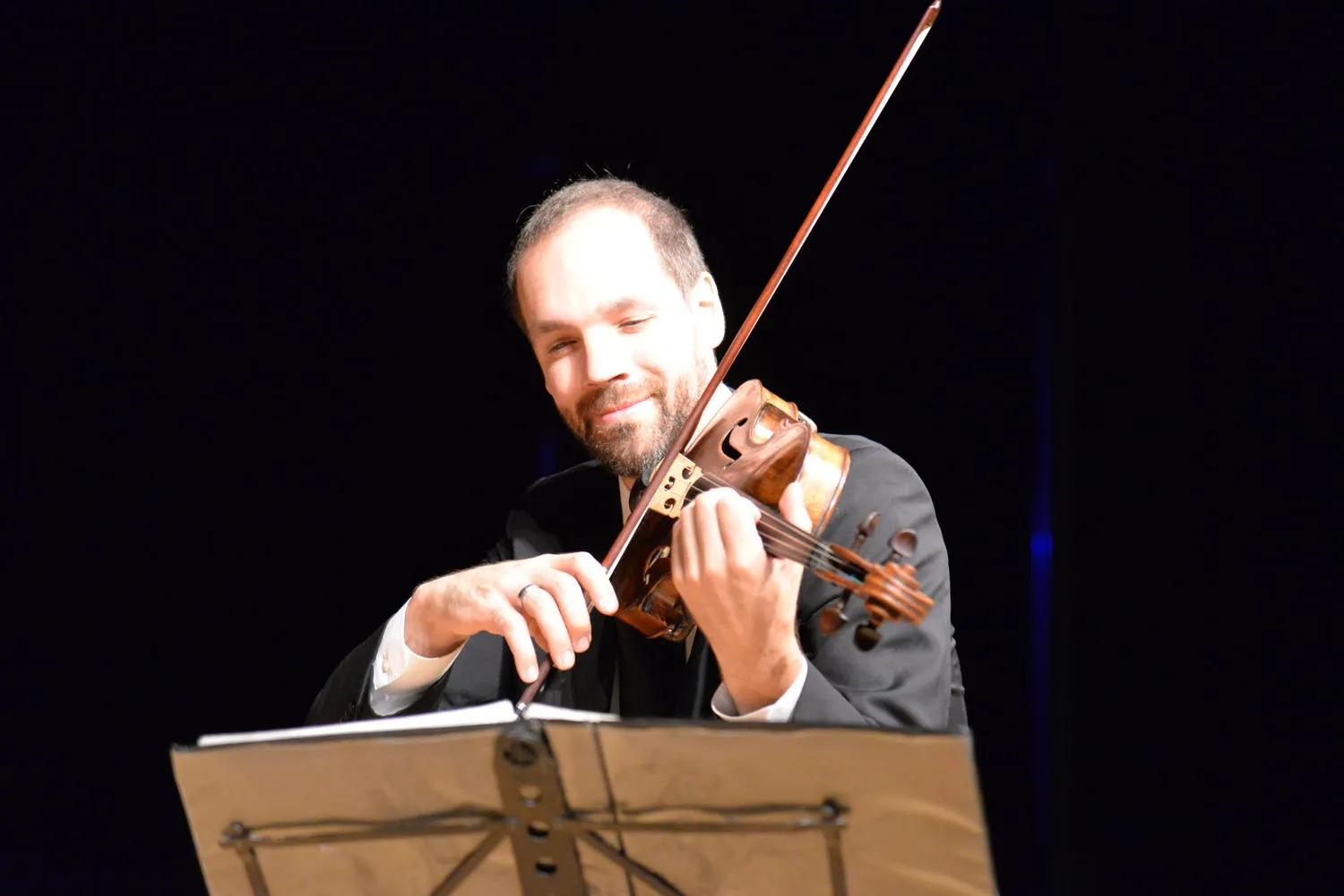 A smiling string player in a dark suit, bow raised above the music stand