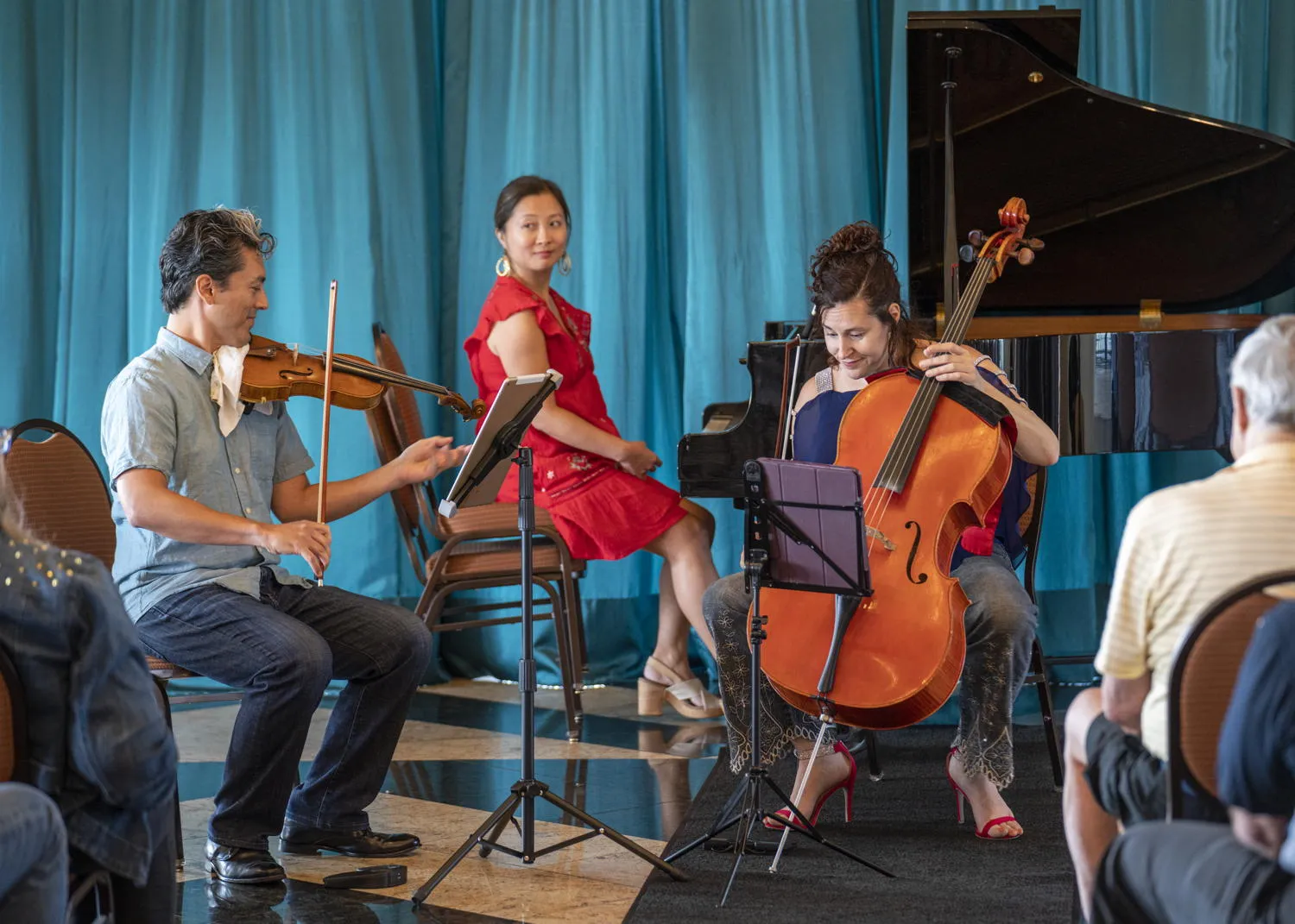 A violinist and a cellist playing as a pianist in red looks on, in front of a teal curtain