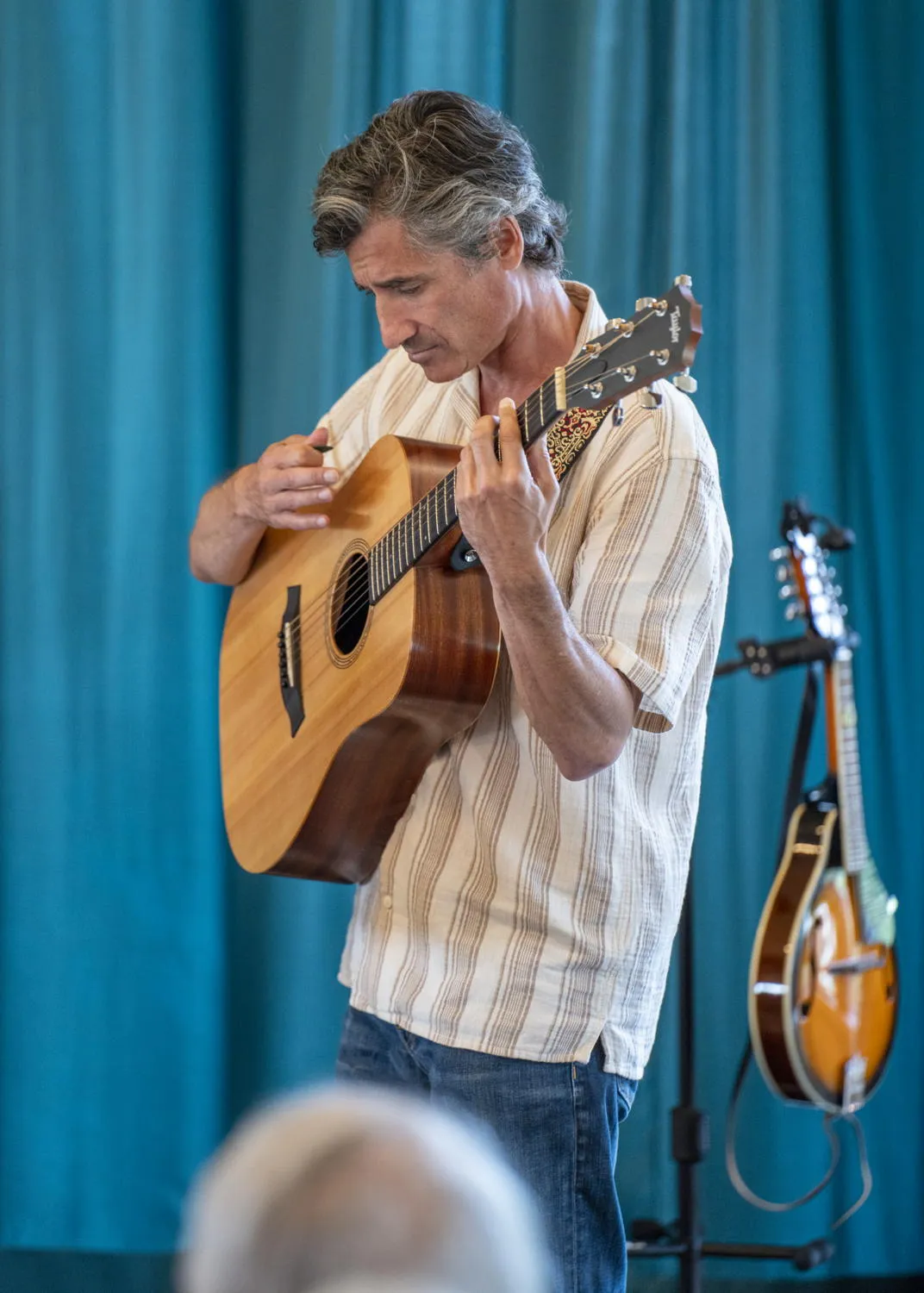 A guitarist playing in front of a teal curtain, with a mandolin on a stand