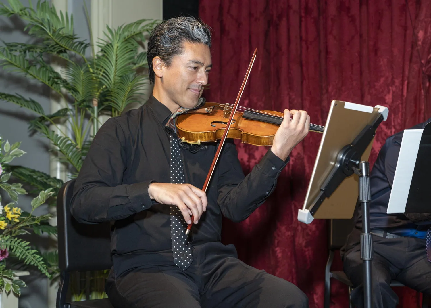 A violinist playing in front of a red curtain and potted palms