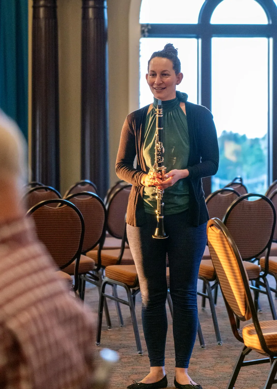 A smiling clarinetist standing among rows of chairs by an arched window