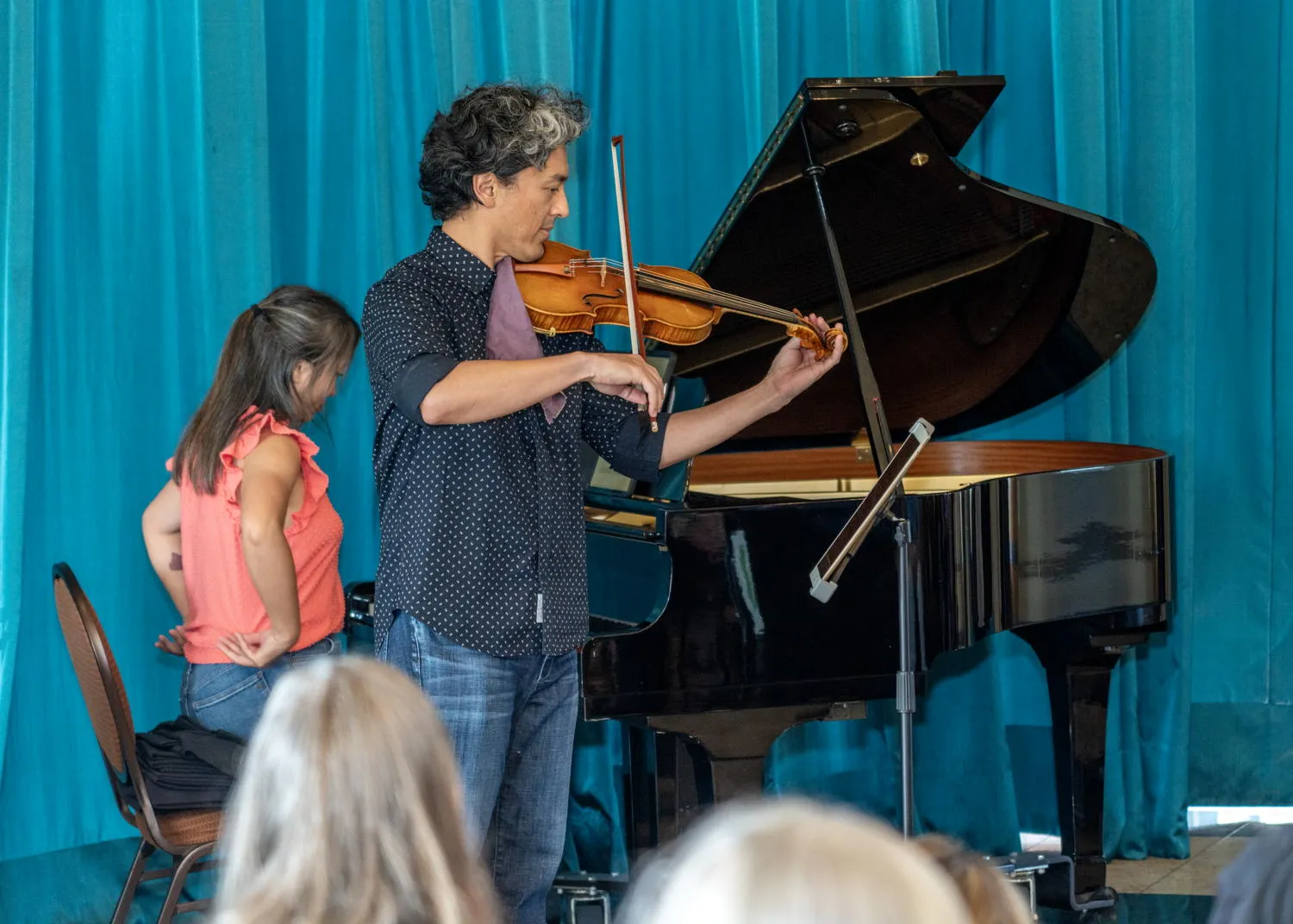 A violinist playing beside a grand piano in front of a teal curtain