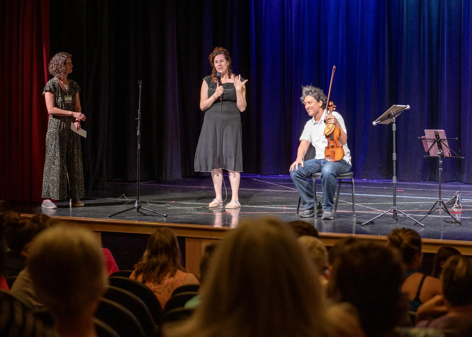 A musician speaking into a microphone on stage while a seated violinist listens