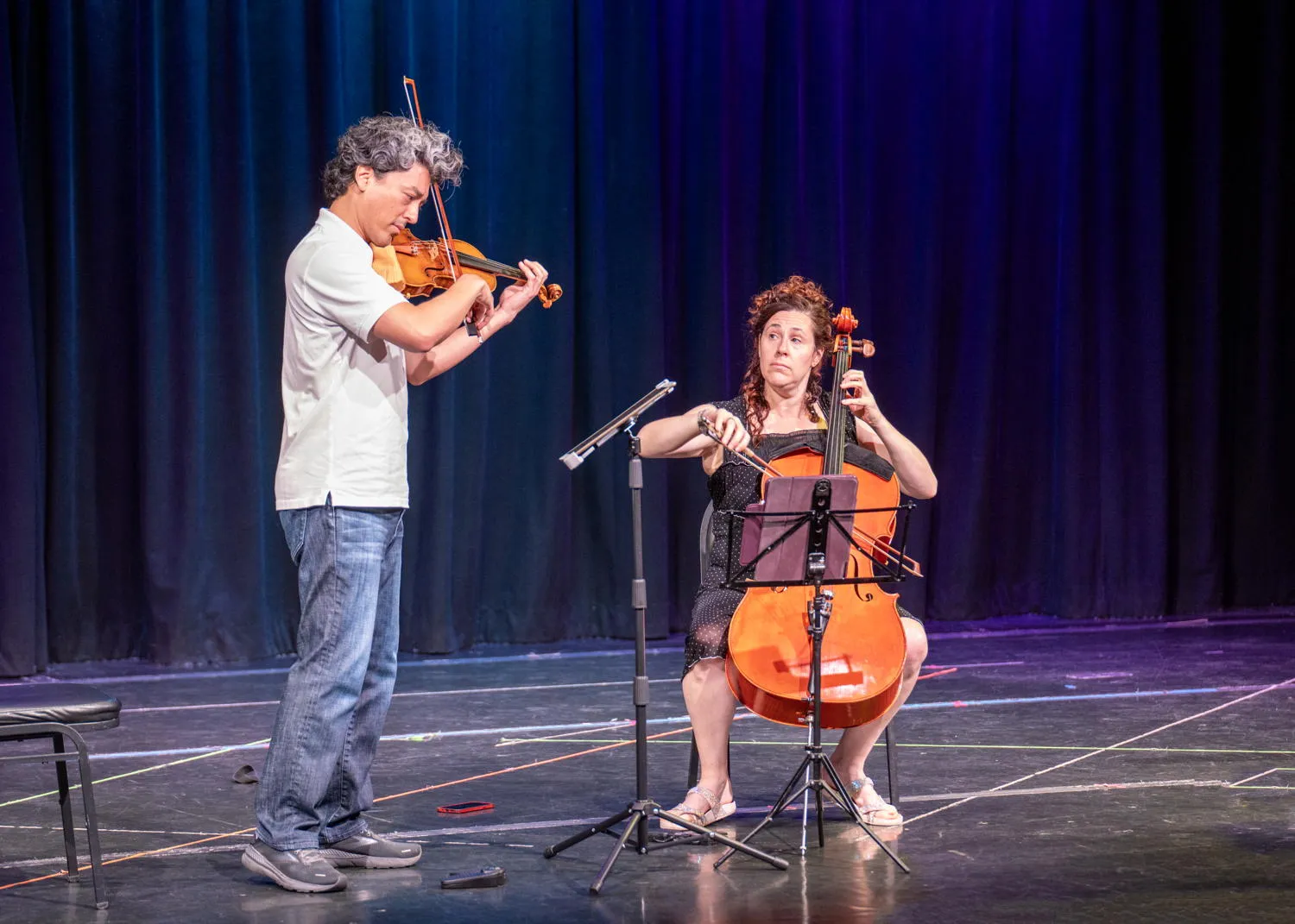 A violinist and a cellist performing on a stage with a blue curtain