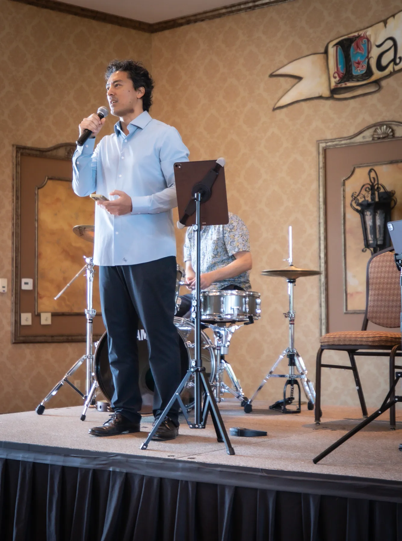 A musician in a light blue shirt speaking to the audience in front of the drum kit